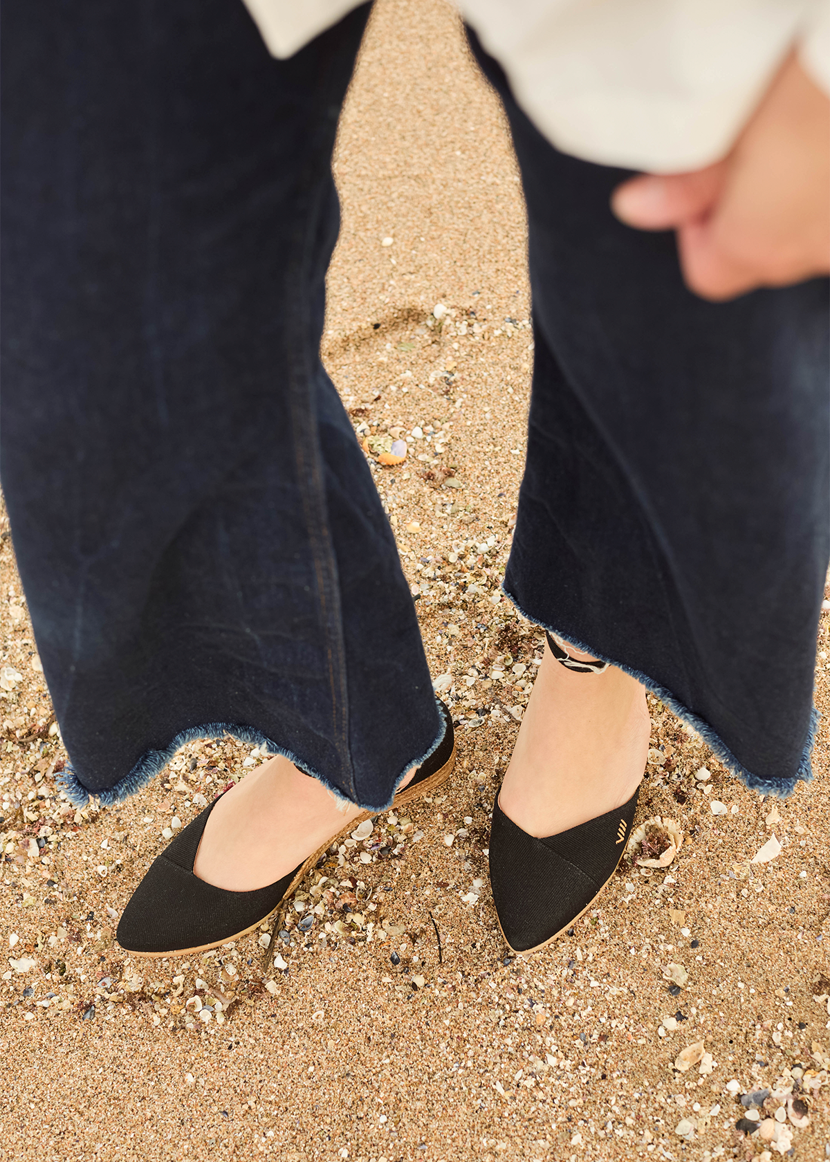 Woman wearing black Montroig canvas flat espadrilles by Viscata on a sandy beach with seashells.
