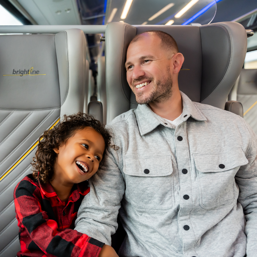 A man and a young child sit together on modern Brightline train seats, smiling and enjoying their time. The child in a red and black checkered shirt leans happily against the man in a light gray button-up jacket.