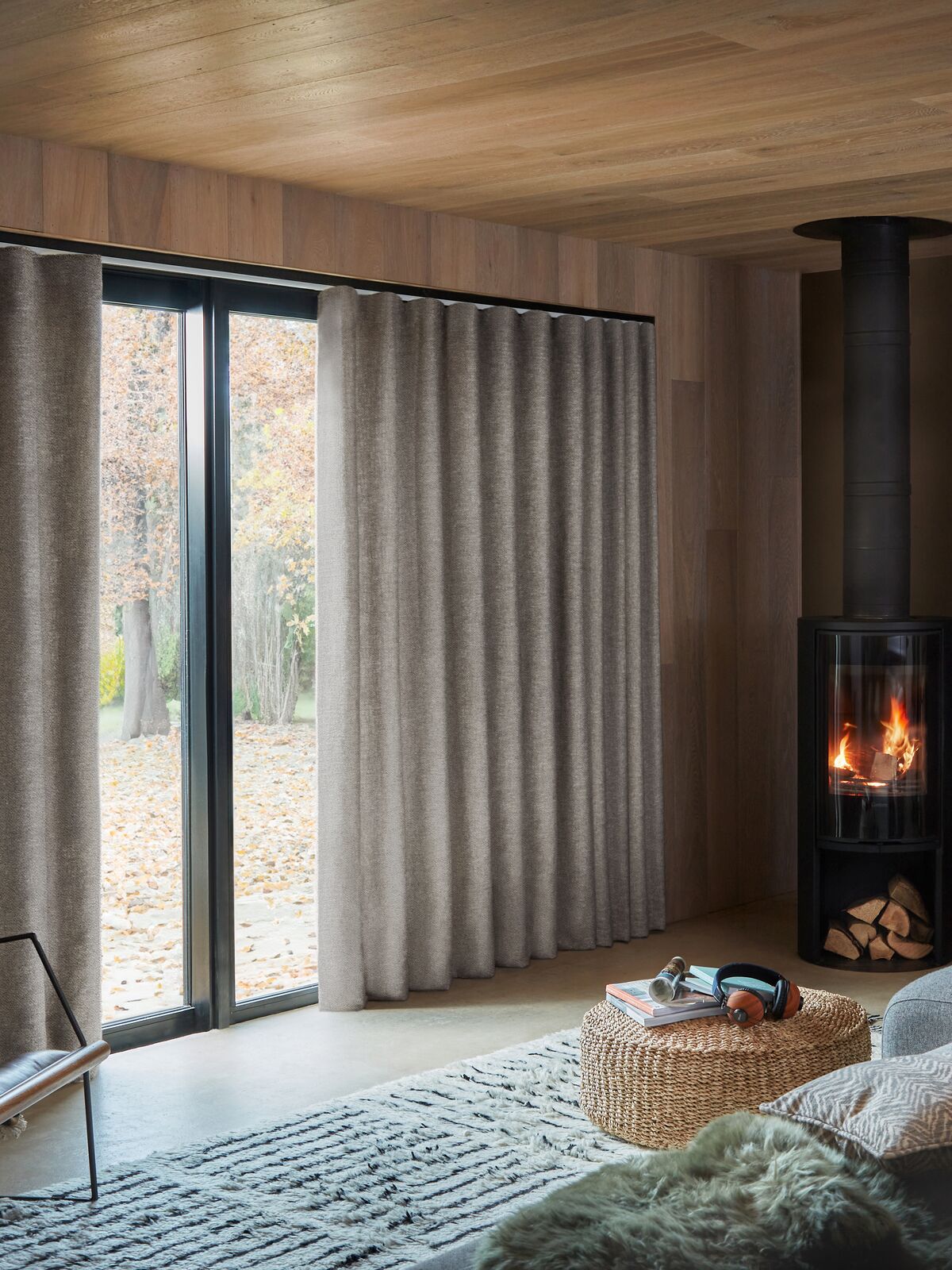 Full-length textured curtains frame sliding patio doors, softly filtering natural light in a cosy living room with a wood-burning stove.
