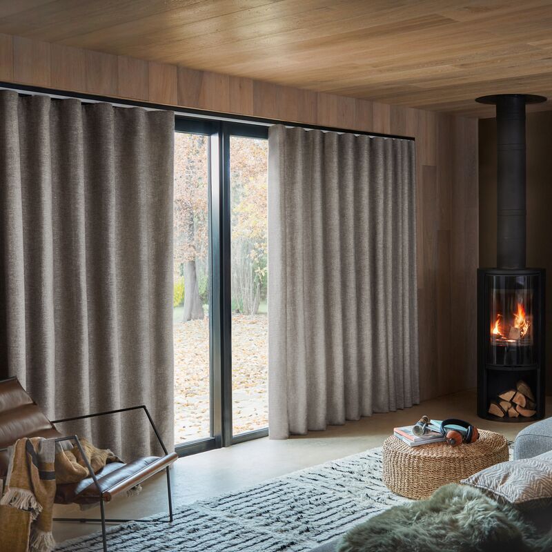 Full-length textured curtains frame sliding patio doors, softly filtering natural light in a cosy living room with a wood-burning stove.