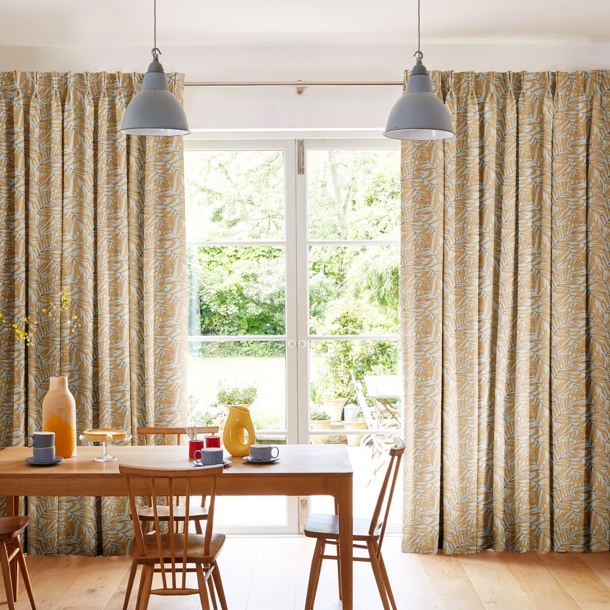 Leaf-patterned curtains frame glazed patio doors, welcoming natural light and garden views into a bright dining space.