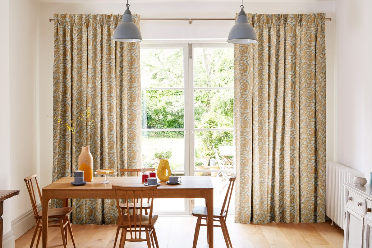 Leaf-patterned curtains frame glazed patio doors, welcoming natural light and garden views into a bright dining space.
