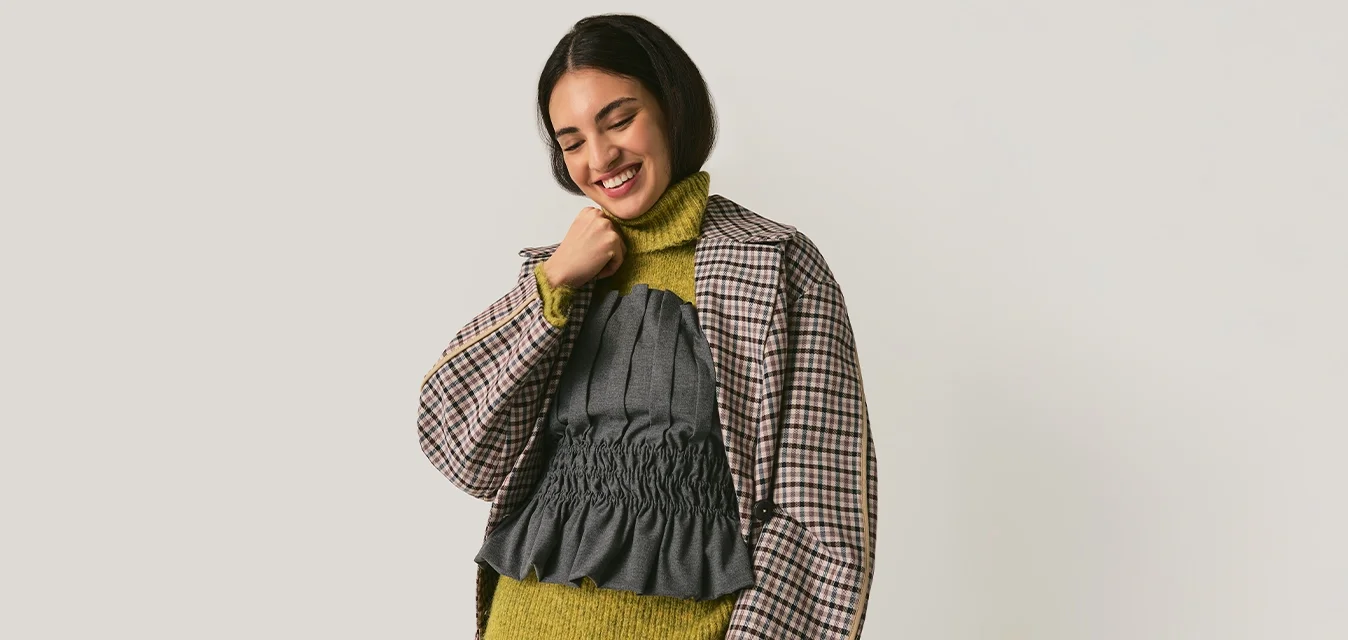 Jonge vrouw die lacht, gekleed in een geruit blazer over een groene coltrui en een gerimpeld grijs topje, met gestreepte grijze broek, staand tegen een effen achtergrond.