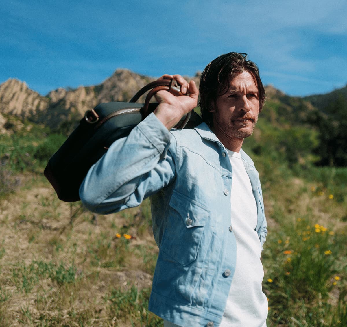 Man in a denim jacket carrying a leather Aviator Brief over his shoulder in a rugged, mountainous landscape.