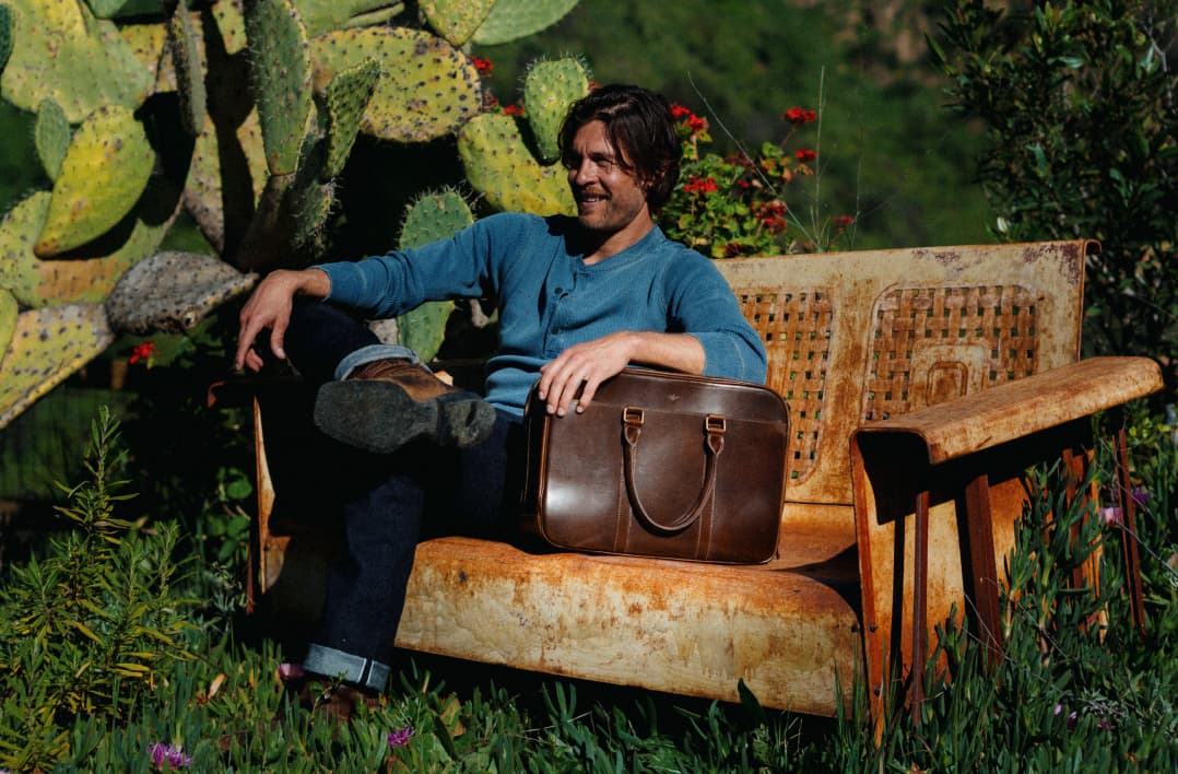 Man walking away carrying a brown leather briefcase overlooking a hillside view, paired with a close-up of the bag resting on a rustic bench beside a seated man's hand.