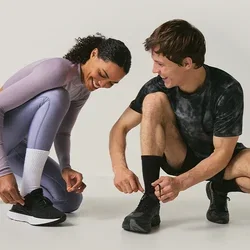 Twee mensen in sportkleding hurken terwijl ze hun hardloopschoenen strikken op een effen, lichtgekleurde achtergrond.