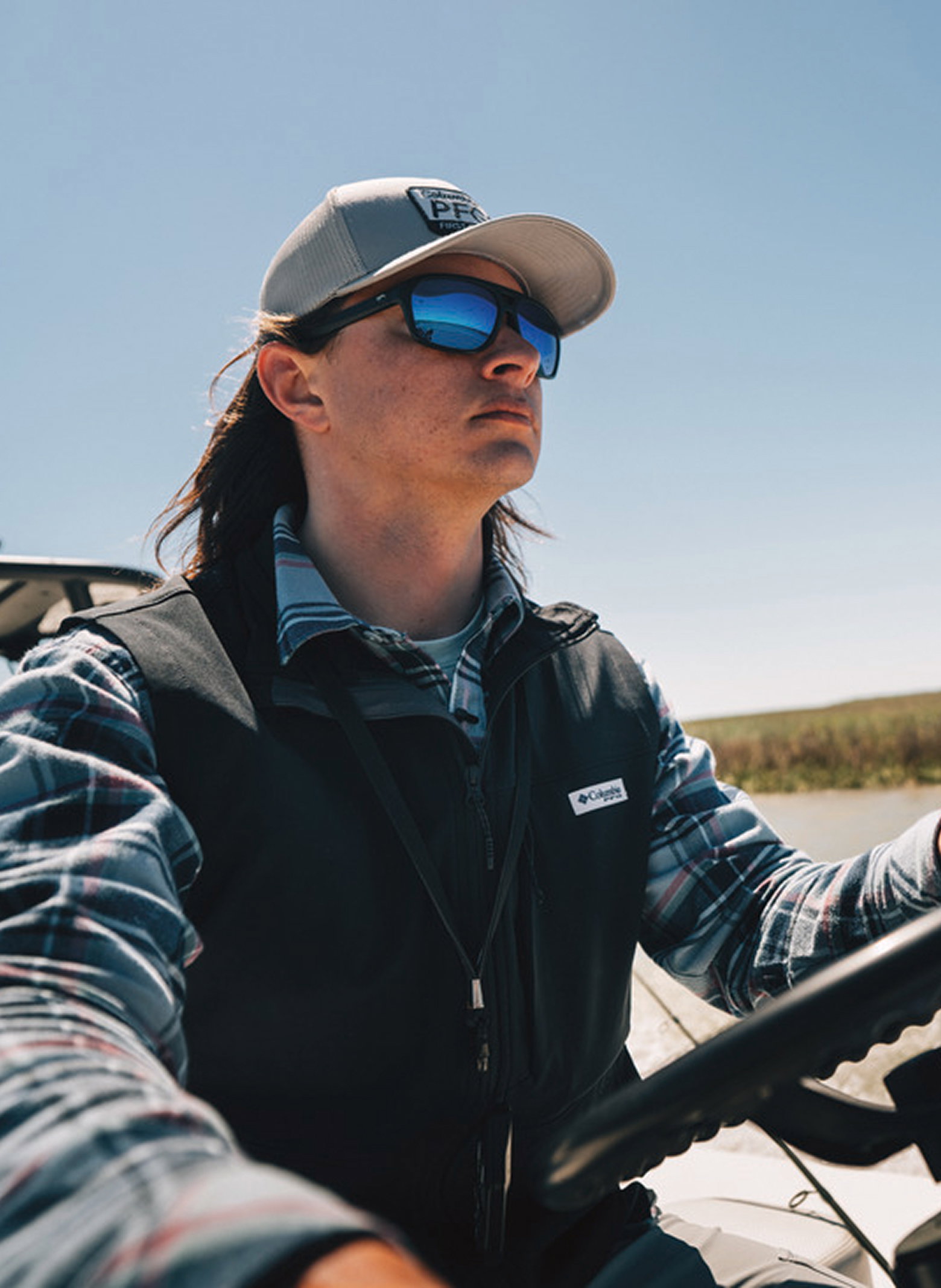 A man driving a skiff, wearing a flannel shirt and vest out in the sun.