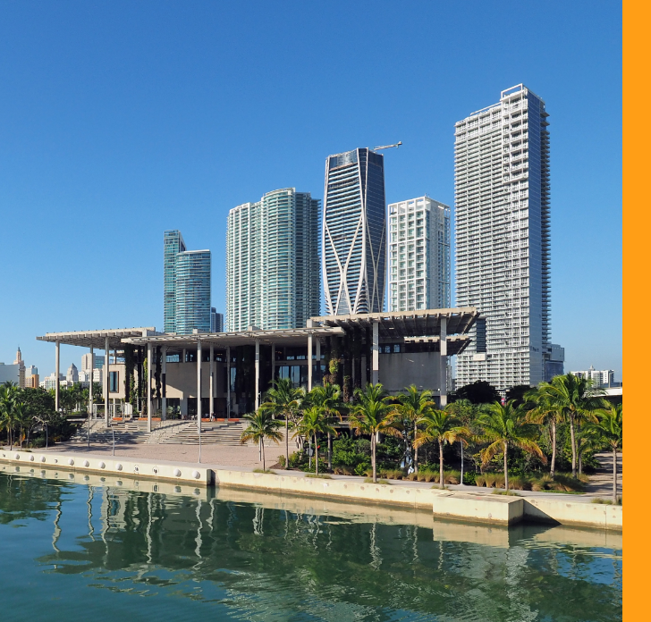  A contemporary waterfront museum stands in the foreground, framed by palm trees and a calm reflective body of water. Behind it rises a cluster of modern high-rise buildings with varied architectural styles and glass facades. The scene is captured under a clear blue sky, giving the image a bright and vibrant atmosphere. Strong vertical lines and geometric forms emphasize the urban character of the cityscape.