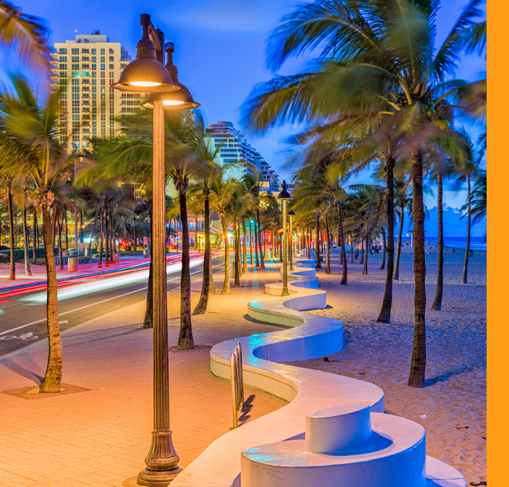 A vibrant coastal promenade is captured at dusk, lined with tall palm trees and modern streetlights. A curving white concrete wall leads the eye along the walkway beside a road with streaking car lights. High-rise buildings glow in the background, adding an urban feel to the tropical beach setting. The sky transitions from deep blue to warm tones, creating a colorful, energetic atmosphere.