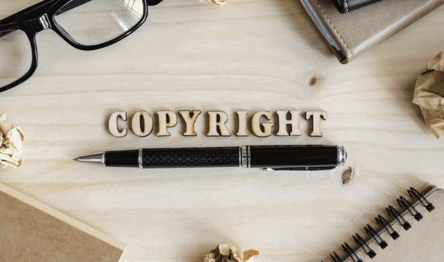Wooden letters spelling "COPYRIGHT" are arranged on a light wooden surface surrounded by glasses, books, crumpled paper, and a pen.