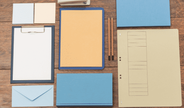 A neatly organized layout of office supplies including a clipboard, folders, envelopes, pencils, a pen, and a stapler, all arranged on a wooden surface.