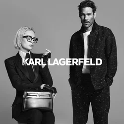Vrouw met kort blond haar en een bril zit op een kruk in een kostuum, met handschoenen en loafers, terwijl ze een handtas vasthoudt; man staat ernaast in een casual donkere outfit.