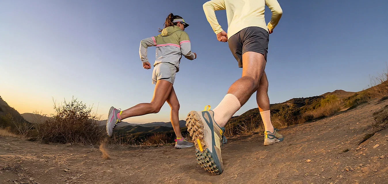 Twee mensen rennen op een onverharde trail in een bergachtig gebied tijdens zonsopgang of zonsondergang, gekleed in sportkleding en hardloopschoenen.