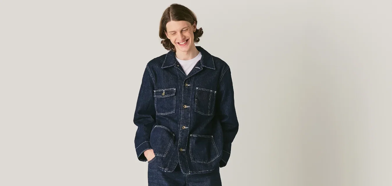 Persoon in een donkerdenim jack en broek, staan met de handen in de zakken tegen een lichtgrijze minimalistische achtergrond.