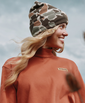A woman wearing an orange sweater and camo beanie against a cloudy sky.