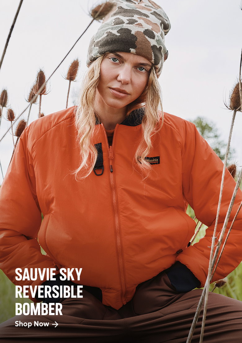 A woman wearing a bright orange jacket and camo beanie with text: SAUVIE SKY REVERSIBLE JACKET