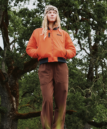 A woman wearing a bright orange jacket and brown pants outside.
