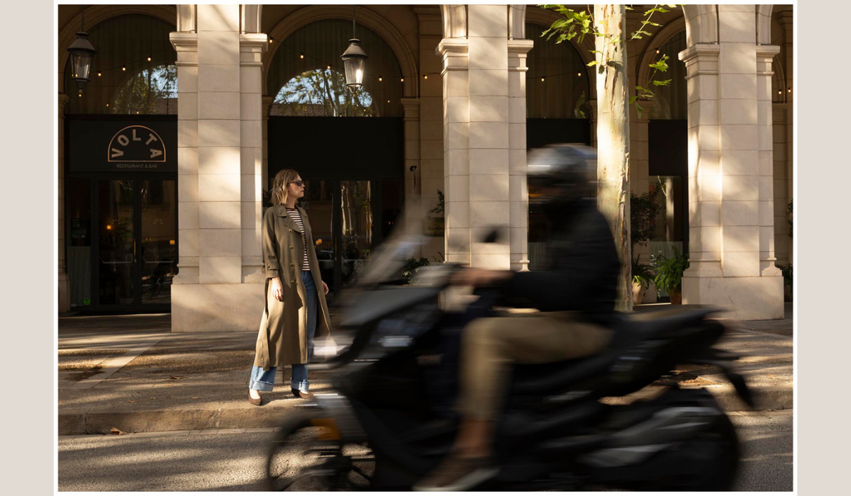 Woman in autumn layers standing on a sunlit city street as a scooter passes by.