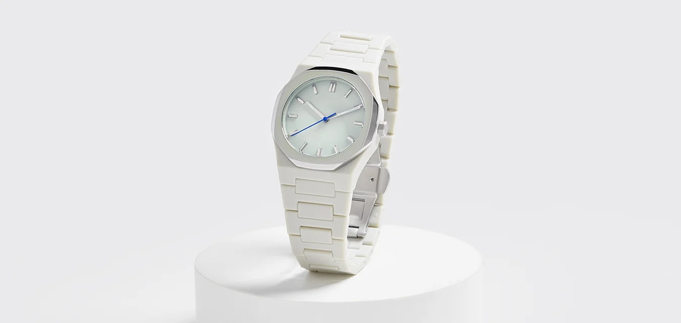 Witte kunststof horloge met een glad, octogonaal gezicht en blauwe wijzers, weergegeven op een ronde witte voet tegen een lichte achtergrond.