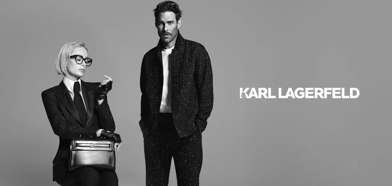 Vrouw met kort blond haar en een bril zit op een kruk in een kostuum, met handschoenen en loafers, terwijl ze een handtas vasthoudt; man staat ernaast in een casual donkere outfit.