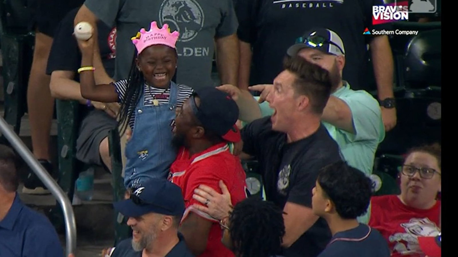 A young fan with a Happy Birthday crown on holds up a baseball as her father holds her