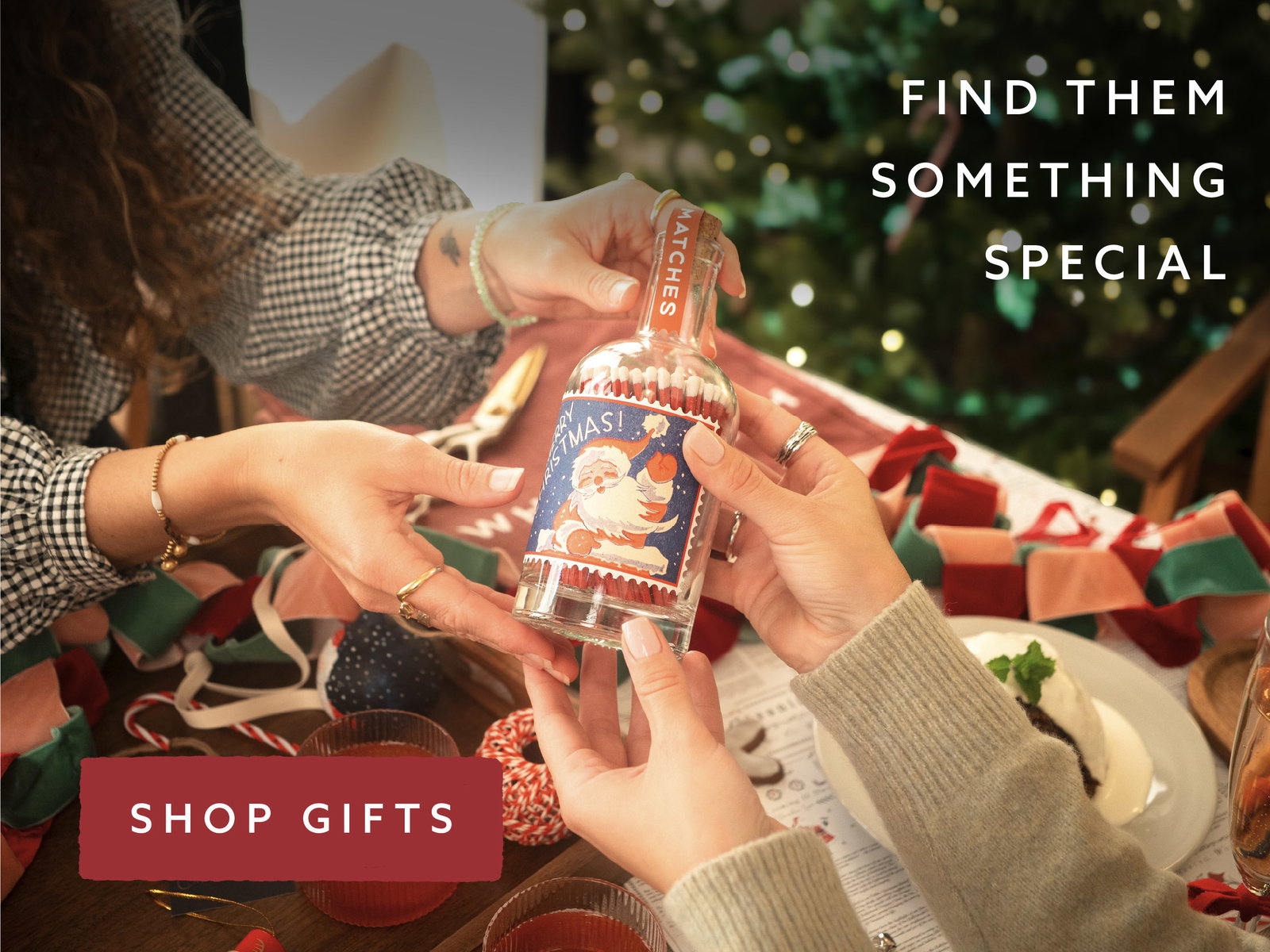 Two people exchanging a festive bottle of decorative matches beside a Christmas tree, with colourful ribbon and gifts on the table. Text reads: “Find them something special. Shop gifts.”