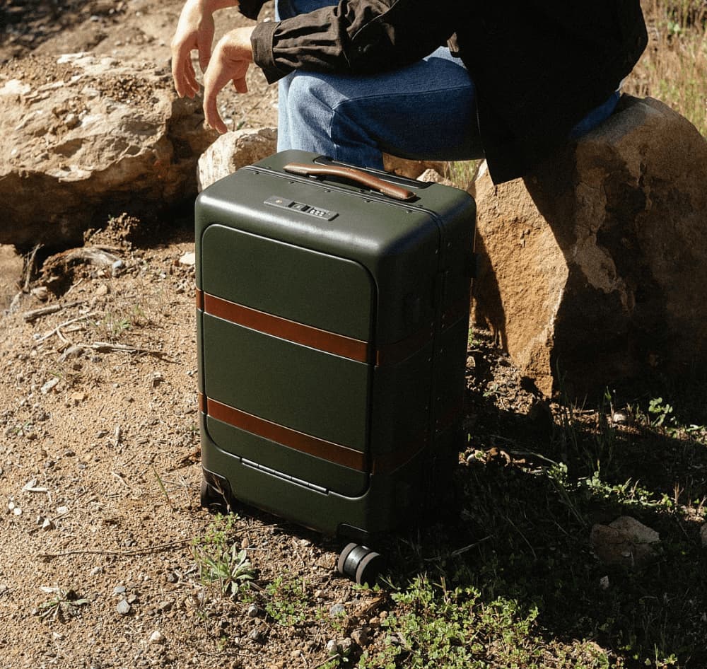 Man in a black shirt and jeans carrying a brown leather briefcase in a grassy mountain landscape, paired with a close-up of the bag hanging on a wooden wall hook.