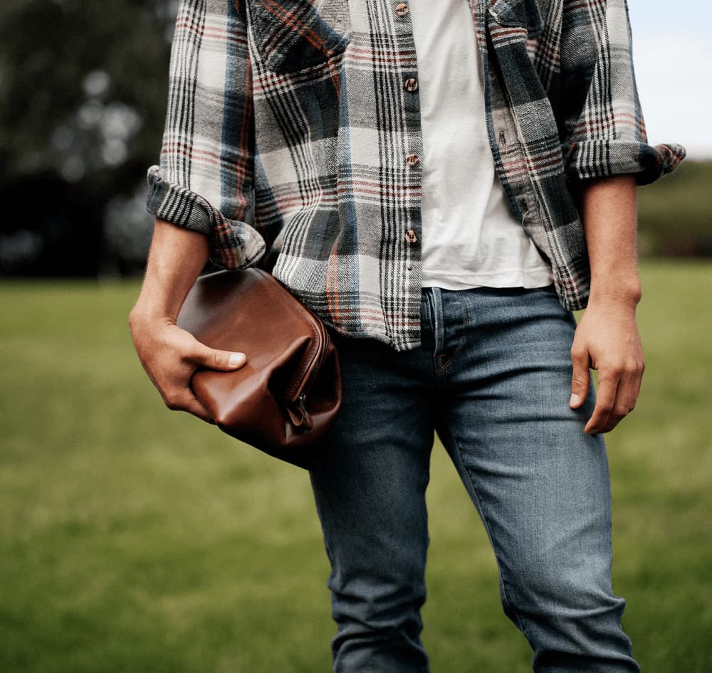 Man walking away carrying a brown leather briefcase overlooking a hillside view, paired with a close-up of the bag resting on a rustic bench beside a seated man's hand.