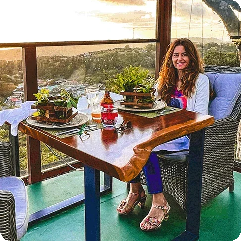 Image Description: Woman seated at a large wooden table smiling at the camera. The table is next to a large pane of glass with a beautiful treetop view just beyond.